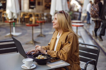 Focused lady working on laptop while sitting at table outdoors with cup of coffee
