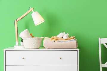 Stack of baby clothes, shoes and glowing lamp on chest of drawers near color wall