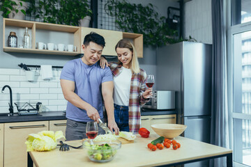 multiracial young happy family, Asian man and woman, rejoice and cook dinner together, cut vegetables for salad, happy couple at home on a date in love