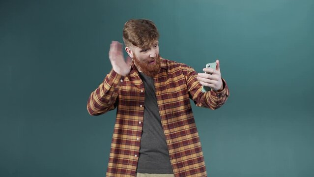 Enthusiastic young redhead man make a video call by phone camera near a green wall