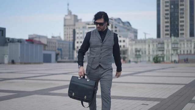 Portrait of serious Caucasian successful businessman strolling on city square with briefcase checking time on watch leaving in slow motion. Confident elegant man in formalwear walking for meeting