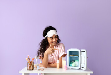 Woman in sleep mask pointing at opened refrigerator on table with cosmetic products