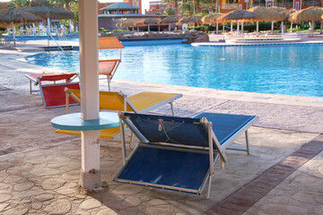Empty deck chairs under straw shade umbrellas on swimming pool side in tropical resort. Summer vacations and getaway concept