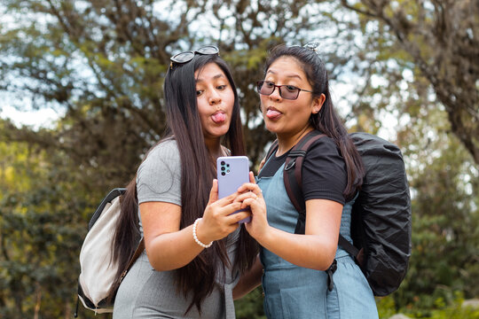 Amigas felices en aire libre,Retrato de dos hermosas chicas mirando el celular mostrando la lengua en un  hermoso parque,Dos chicas usando el telefono en medio de un camino rodeado de monta&ntilde;a