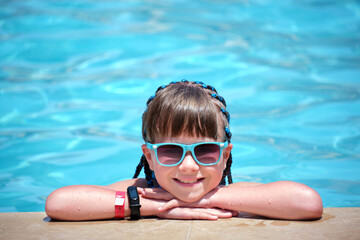 Young joyful child girl resting on swimming pool side with clear blue water on sunny summer day. Tropical vacations concept