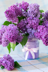 Vertical photo of a bouquet of flowers with lilac petals and green leaves in a vase with a bow on a table with a checkered tablecloth