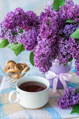 Vertical photo of a bouquet of flowers with lilac petals in a vase with a bow on a table with a checkered tablecloth next to a white mug of tea and a vase with sushki