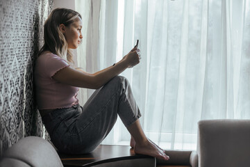 A girl with long hair is sad and reads something on the phone, experiences negative emotions, is upset, sad. Against the background of the window of the house, during the day