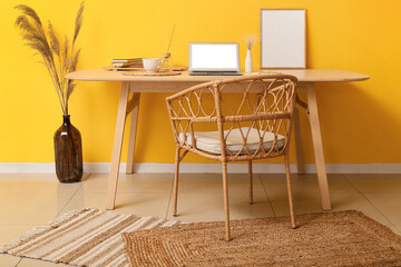 Modern workplace with laptop, cup of coffee, frame and pampas grass in vase near yellow wall
