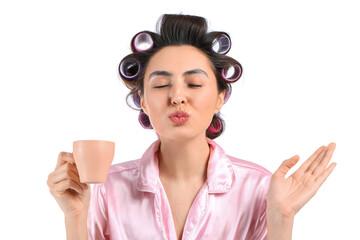 Beautiful young woman in pajamas and hair curlers drinking coffee on white background
