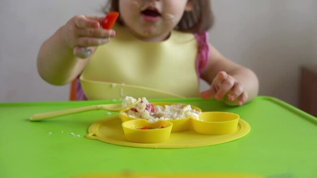 Close up of lovely baby girl eating have breakfast eggs vegetables sausage by herself at home. Adorable possitive happy toddler child eats with dirty face at table indoor. Food, nutrition healthy life