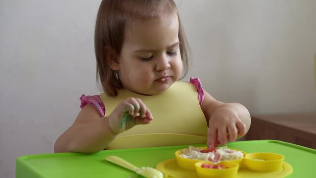 Close up of lovely baby girl eating have breakfast eggs vegetables sausage by herself at home. Adorable possitive happy toddler child eats with dirty face at table indoor. Food, nutrition healthy life