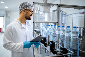 Technologist in sterile uniform and hairnet checking production of water in bottling factory.