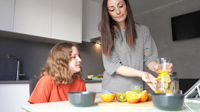 Happy brunette mom with her teenage daughter preparing healthy vegan breakfast in the kitchen at home. Orange juice and lemon water, citrus as a source of energy and vitamins for breakfast. 