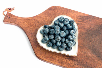 Fresh juicy delicious blueberries lie on a white original plate in the form of a heart, a kitchen wooden board. White background.