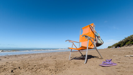 Chair on the beach on the sand, beach bag and flip flops. Blue sky and copy space