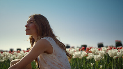 Thoughtful girl sitting in flower field in sunlight. Woman dreaming among tulips
