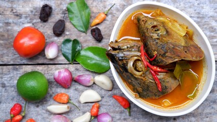 Asam pedas kepala tongkol is Indonesian sour and spicy fish head curry on a wooden background and some spices in the side 