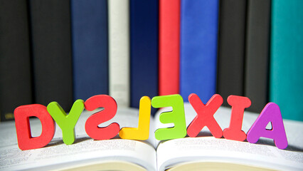 Bright colorful wooden toy blocks on an open book spelling dyslexia. Learning challenges disabilities. S, L and E are backwards. Hard bound books lined up in background