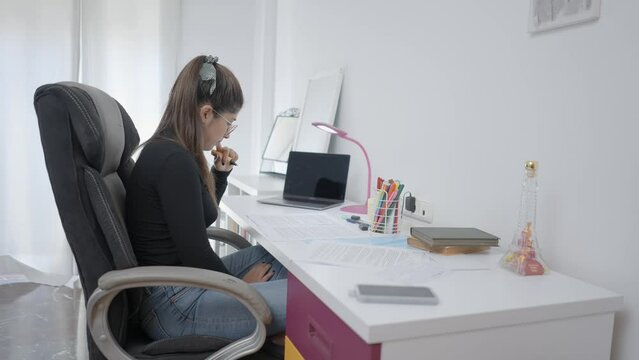 Young colombian girl student studying at home reading compendium notes in exercise book using laptop computer for online self education doing creative coursework research homework assignment at desk.