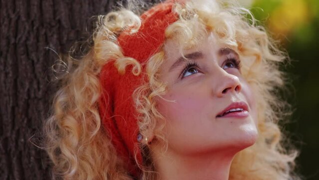 Face of Young Blonde Woman With Red Headband in Curly Hair Sitting Under Tree on Sunny Autumn Day, Close Up