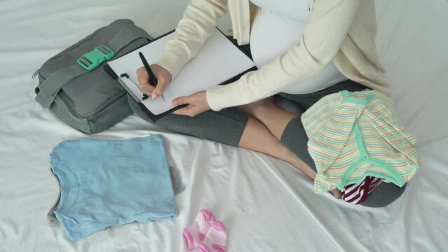 A pregnant woman write down on paper for recheck necessary for newborn.  Mother is folding and keep clothes to prepare for the upcoming baby. Happy for new born concept...
