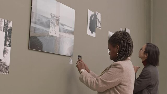 Medium slowmo shot of two diverse visitors looking at photo exhibition in modern art gallery. African-American woman scanning photo qr-code with smartphone to find more info about it