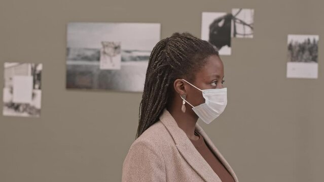 Waist-up slowmo portrait of young African-American woman in face mask looking at camera standing at contemporary art gallery with abstract black-and-white photos hanging on wall in background