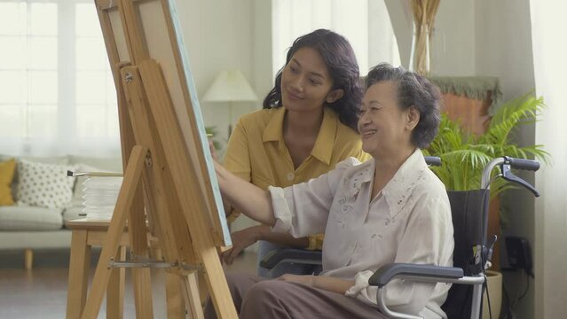 Female teacher teaching senior woman to paint picture at living room. Senior woman sitting wheelchair while paint picture. Healthcare, Lifestyle and Hobby concept.