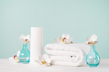 Fototapeta na wymiar A folded white towel, a bottle of shampoo or shower gel and orchid flowers on a light blue background. Bath mockup with copy space.