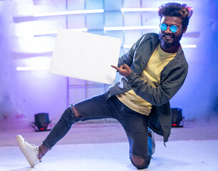 young smiling hip hop dancer showing white empty board or placard by looking at camera on stage - concept of advertisement, ads promotion and professional dancer