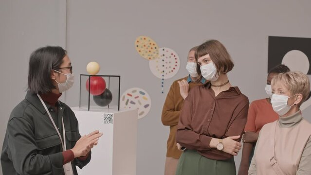 Medium slowmo shot of diverse group of tourists in face masks visiting exhibition at museum of contemporary arts having conversation with young Asian male guide