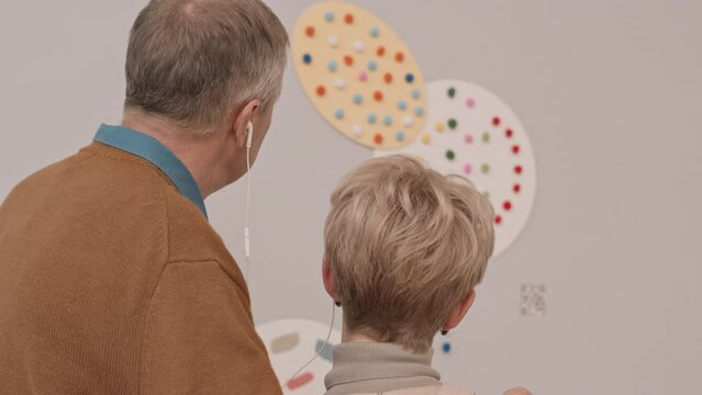 Rear-view slowmo shot of mature Caucasian couple in headphones looking at art object on wall in modern art museum and listening audio guide