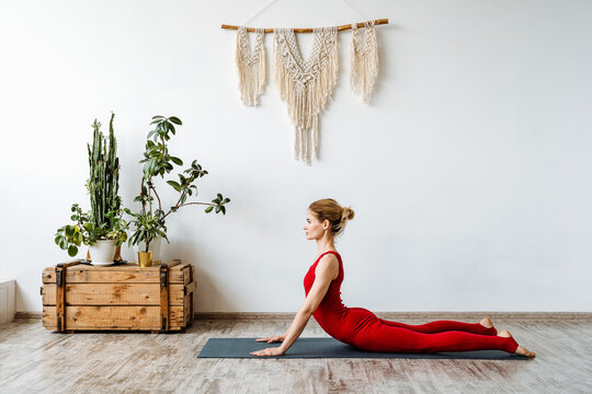 Bhujangasana cobra pose. The girl practices yoga on the mat at home. A young woman practices hatha yoga against the backdrop of the boho interior of an apartment. Red sportswear.