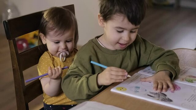 Brother playing with babysister while enjoying leisure time together at home drawing with colored pencils while sitting at table. Children education. Happy face