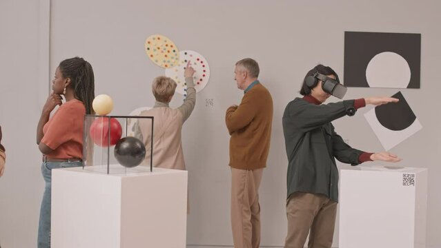 Medium slowmo shot of diverse group of people examining conceptual art objects during exhibition in contemporary art museum. Young man wearing vr headset