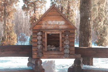 A big wooden birdhouse reminding a wooden rural house made with timber. During a snowy winter or spring in the woods of an urban park