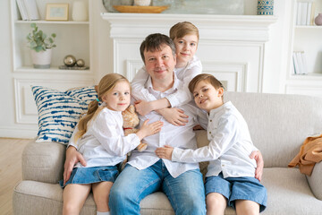 Happy caucasian family at home, cute son and daughter hugging dad, father fooling around with little son and daughter, siblings enjoying active time together sitting on sofa in living room