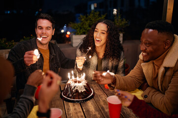 Group of diverse young adults sitting at boho themed rooftop birthday party lighting sparklers from...