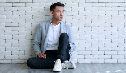 Portrait shot of Asian young cool smart handsome muscular undercut hairstyle male businessman in casual gray suit jacket sitting holding hands at chin look at camera leaning on brick wall background