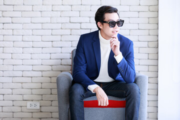 Studio shot Asian young smart handsome successful professional male businessman in casual blue suit turtle neck shirt sunglasses sit on armchair looking at camera on brick wall background