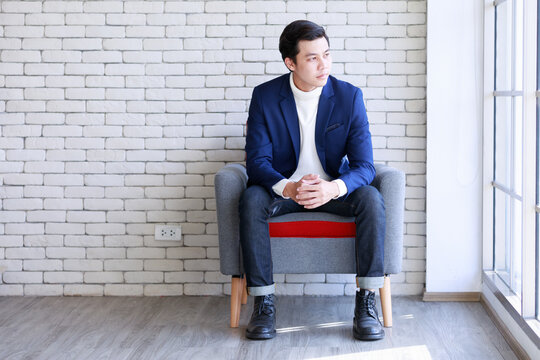 Studio Shot Asian Young Smart Handsome Successful Professional Male Businessman In Casual Blue Suit Turtle Neck Shirt Sit On Armchair Holding Hands Together Looking Outside On Brick Wall Background