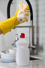  White bottle with dishwashing gel against the background of a sink and pouring foam from a sponge.