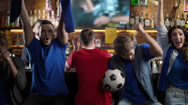 International football fans watching a football match at the bar and enjoy scoring a goal, fan of the losing team is sitting at the bar.
