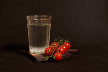 A faceted glass of vodka and a branch of cherry tomatoes on a wooden stand