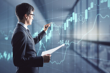 Attractive young european businessman with glowing candlestick forex chart grid hologram standing in blurry office interior. Innovation, trade and finance concept. Double exposure.