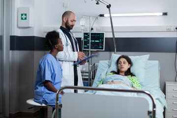 Obraz premium Recovering woman on hospital bed with iv drip line and vital functios monitored being consulted by doctor with medical history on clipboard. medic doing consult with nurse looking at patient chart.