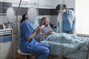 Nurse sitting at patient bedside writing medical observations on clipboard for middle aged man recovering after surgical intervention. Doctor adjusting intravenous medication iv drip line flow. - Powered by Adobe