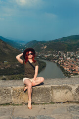 A young tourist girl on the background of a stunning view point of the Jvari Monastery. Mtkvari river meeting the Aragvi river, Summer day. Mtskheta. Georgia. Vertical photo.