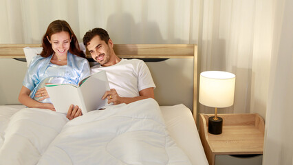 Happy pregnant wife holding belly and relax on bed beside beloved husband and enjoy watching entertainment media on book together with smile and laugh